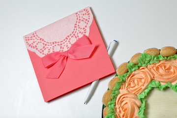 Sponge cake with cream cheese, decorated with Savoiardi cookies. On the side, wrapped in a ribbon tied to a bow. Near a greeting card and pen.