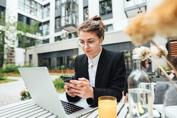 Woman sitting in cafe and working on laptop. Businesswoman wearing glasses at cafe with a computer typing text on his smartphone