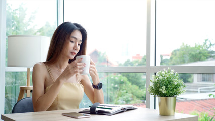 Fototapeta premium Young beautiful Asian woman enjoying a relaxing moment working and using mobile phone as mobile smart payment to pay for a coffee in the coffeeshop on a bright sunny day