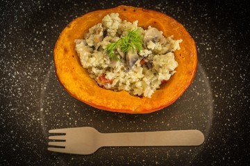 Top view of half of a baked orange pumpkin stuffed with millet grains and mushrooms, a healthy homemade vegetarian dinner on black background with brown wooden fork.
