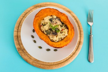 Top view of half of baked orange pumpkin stuffed with millet grains and mushrooms, a healthy homemade vegetarian dinner served on white plate with seeds. Wooden chopping board and silver fork on turqu