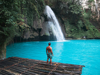 Fit man alone on the bamboo raft in front of the waterfall with turquoise water in Kawasan Falls in...