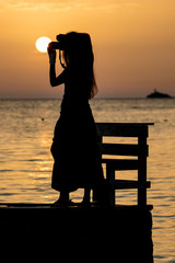 Beautiful female silhouette of unrecognizable slim tall woman with camera taking photos near bench at sunset in front of golden Ionian Sea water near beach. Dusk as seen from Ksamil, Albania, spring