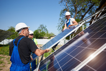Team of three professional workers installing heavy solar photo voltaic panels on high steel platform. Exterior solar system installation, alternative renewable green energy generation concept.