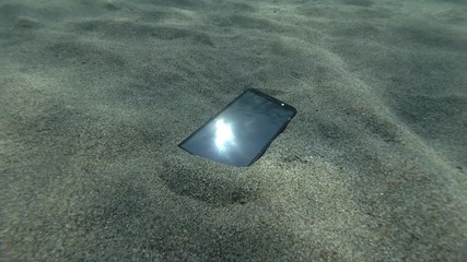 Smartphone lies on a caved seabed on its screen reflects the sun with rays and surface of blue water with small waves. Underwater view. Mediterranean Sea, Europe.