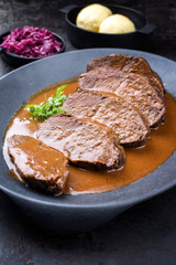 Traditioell geschmorter marinierter deutscher Sauerbraten vom Rind mit Blaukraut und Kartoffel Knödel in würziger brauner Sauce als closeup in einem Modern Design Gusseisen Teller