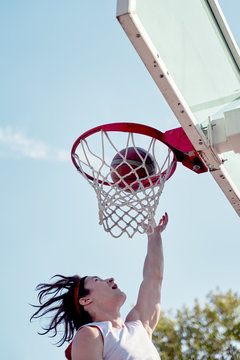 Photo of young athlete man throwing ball into basketball hoop on sports field on street on summer day.