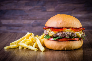 Craft beef burger with cheese, italian peperoni, tomato, basil leafs and french fries on wood table and rustic background.