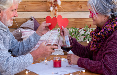 Happy senior couple show two red heart shapes. Sitting outdoor in a romantic corner made by wood. Yellow lights. Two red wineglasses to celebrate a positive moment
