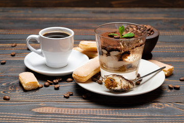 Portion of Classic tiramisu dessert in a glass, cup of coffee and savoiardi cookies on wooden background