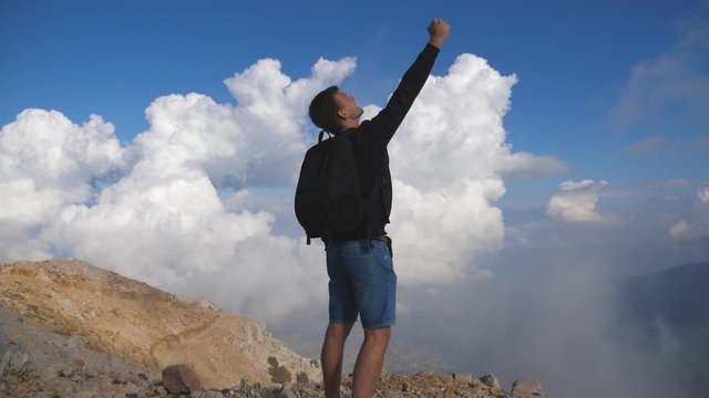 Young male hiker with backpack reaching up top of mountain and raising hand up. Boy tourist standing on the edge of the hill and victoriously outstretching arm. Guy enjoying freedom. Slow motion