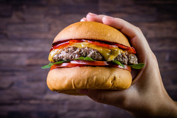 Hand holding a craft beef burger with cheese, italian peperoni, tomato and basil leafs on rustic...