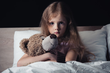 scared kid embracing teddy bear while sitting under blanket and looking at camera isolated on black