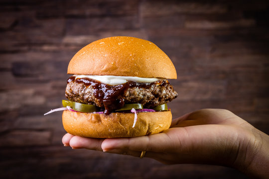 Hand holding a craft beef burger with cream cheese, purple onion, jalapeno pepper on wood table and rustic background.