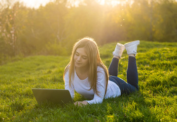 Young beautiful woman with a laptop lies on the grass in sunset.