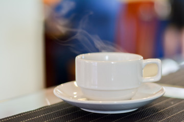 Hot coffee and tea in white ceramic cup on plate with smoke in blurry background.