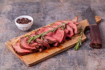 Slices of medium rare roast beef meat on wooden cutting board, old knife, pepper and rosemary twigs on gray marble background. Gourmet food. Raw meat beef steak.