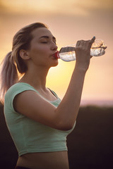 silhouette profile of a beautiful girl drinking water after running a distance in a field at sunset, athletic woman with bottle outdoors concept sport and healthy lifestyle