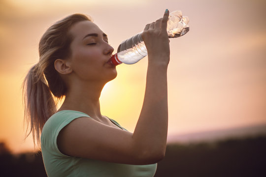 silhouette profile of a beautiful girl drinking water after running a distance in a field at sunset, athletic woman with bottle outdoors concept sport and healthy lifestyle