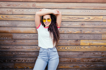 Stylish beautiful young hipster girl in jeans or casual wear yellow glasses posing on backdrop of wooden wall. Concept of style and trends. Copyspace. Glad lady with curly hairstyle, happily emotions