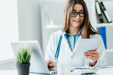 Smiling young female doctor working at the clinic , she is using a computer and writing medical reports.