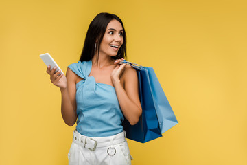 excited woman holding smartphone with shopping bags isolated on orange