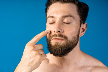 Obraz premium Cropped portrait of handsome, attractive man smearing foam for shave on his face looking at camera isolated on blue background