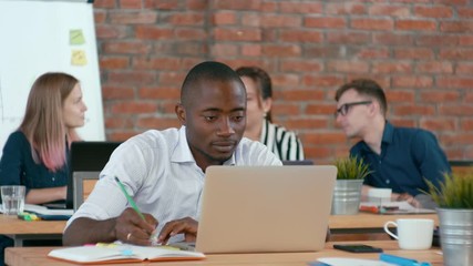 Young African Man is Working on Laptop and Writing by Pencil in Planner in Authentic Loft Office with Diverse Multiethnic Team. Students Discuss Project. 4K Medium Shot