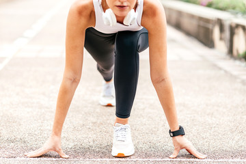 Cropped portrait of motivated woman getting ready to start running on stadium. Young sportswoman with headphones and smartwatch going to run outdoors. Summer training concept. Horizontal shot