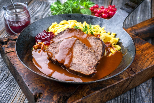 Traditioneller geschmorter marinierter deutscher Sauerbraten vom Rind mit Sp&auml;tzle und Preiselbeer Marmelade in in w&uuml;rziger brauner Sauce  als closeup in einer rustikalen gu&szlig;eisernen Bratpfanne