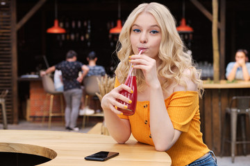 Portrait of pretty blond girl looking in camera while drinking lemonade in courtyard of cafe