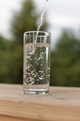 A jet of water flowing into a transparent glass with water and air bubbles standing on a wooden board.