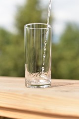 A stream of water flowing into an empty transparent glass standing on a wooden board.