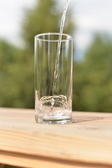 A stream of water flowing into an empty transparent glass standing on a wooden board.