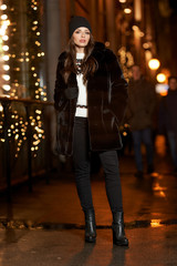 Outdoor night portrait of young beautiful smiling woman standing and posing at street with festive decoration. Christmas decorated city. Female model in striped black and brown fur coat.