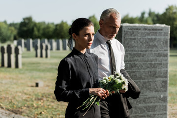 bearded man with grey hair walking near woman with flowers on funeral