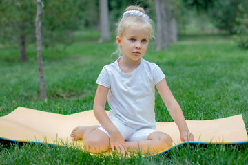 A little disgruntle blue-eyed, curly blonde girl in a black and red gymnastic suit is doing stretching on the blue yoga carpet in the park