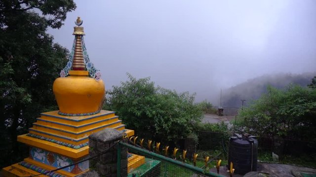 Panning shot of a bhuddhist temple roof with gold spires, hidden behind a green fence showing the clouds, fog and beautiful hill slopes of McLeodganj