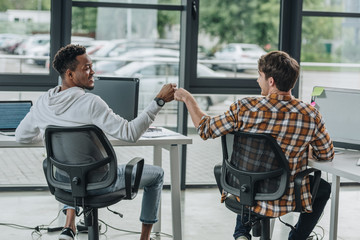 Young multicultural programmers doing fist bump while sitting in office together