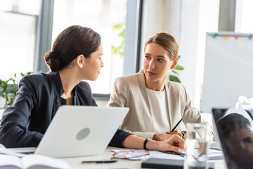 two businesswomen in formal wear at table looking at each other during conference in office