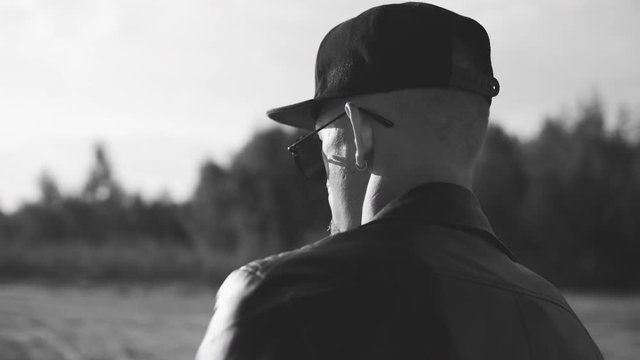Portrait shot of young man walking on the road. View from the back. Caucasian male dancer in sunglasses. Tough guy in black leather jacket. Following shot of the traveling man. Black and white version