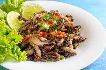 Grilled squid topped with seafood sauce on a white plate with  lettuce and lemon decoration placed on a blue wooden table.