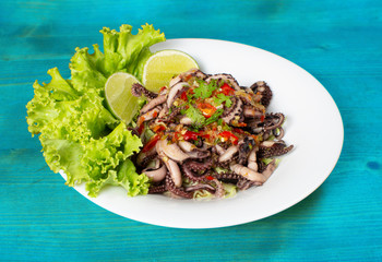 Grilled squid topped with seafood sauce on a white plate with  lettuce and lemon decoration placed on a blue wooden table.