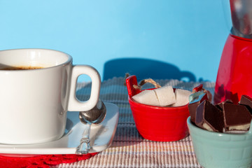 Cup of hot coffee/tea with sugar bowl and chocolate on blue background in sunny morning