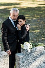 selective focus of senior man hugging upset woman on graveyard