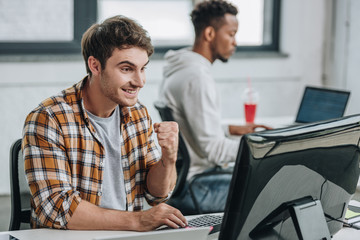 Fototapeta premium cheerful programmer showing winner gesture while working near african american colleague