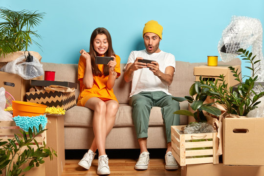 Photo of addicted teenagers focused in smartphones, obsessed with playing video games, moves in new apartment for living, many boxes with personal stuff. Real estate, home and people concept