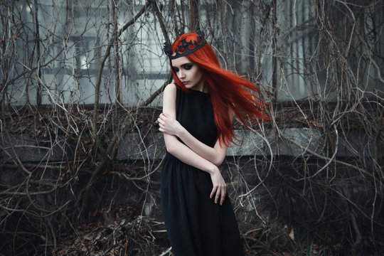 A fragile sad goth girl with red hair in a crown and a black dress stands in the middle of a gloomy landscape