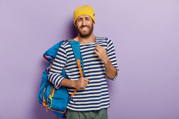 Photo of displeased European man with upset expression, clenches teeth nervously, carries blue rucksack, travels in different countries, wears yellow hat and stirped sweater, poses over purple wall