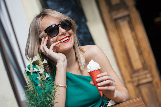 attractive blonde young woman in sunglasses, green dress and with red lipstick sits at table of street cafe. Girl holding a red ice cream cone and talking on mobile phone